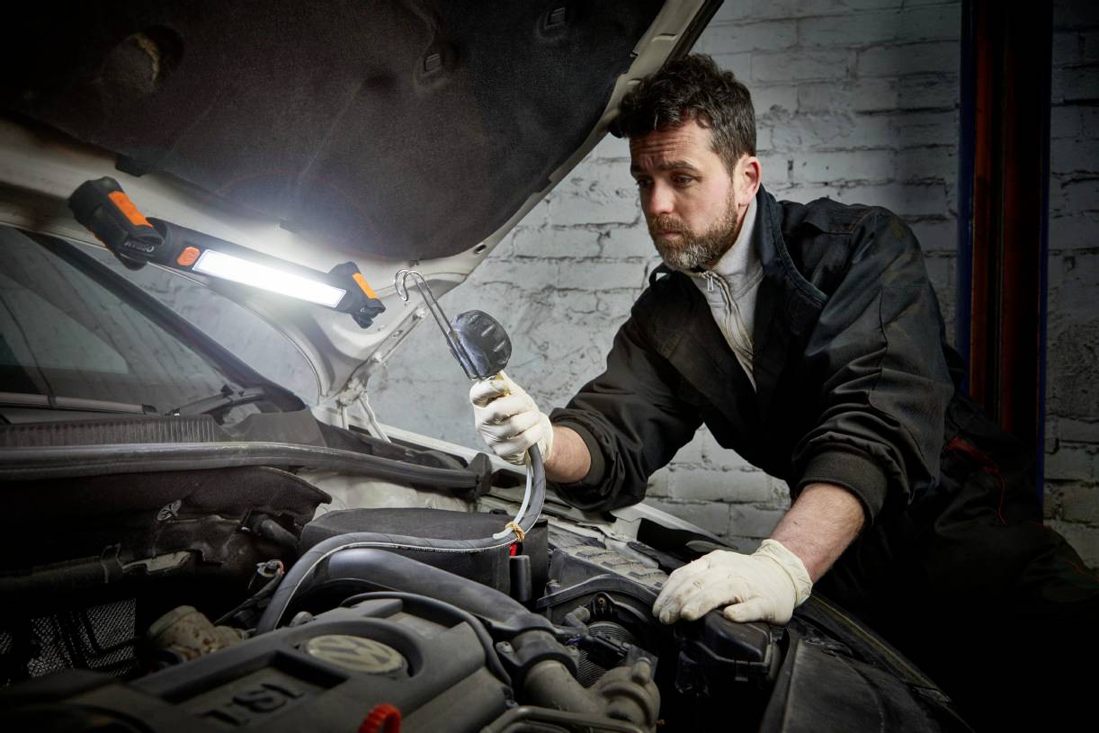 Mechanik w kombinezonie sprawdza silnik samochodu w warsztacie. Trzyma urządzenie diagnostyczne, podczas gdy światło robocze oświetla scenę.