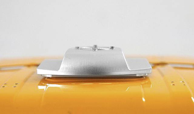 Srebrne urządzenie o prostokątnym kształcie na błyszczącej, pomarańczowej powierzchni, prawdopodobnie będące rozwiązaniem montażowym.