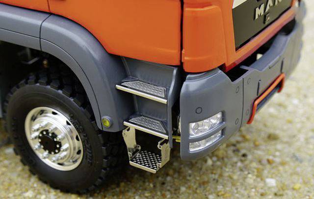 Zbliżenie pomarańczowej ciężarówki z wyraźnie widocznymi stopniami wejściowymi i oponami na żwirowej nawierzchni. Ostrość zdjęcia skupiona jest na szczegółach stopni wejściowych.