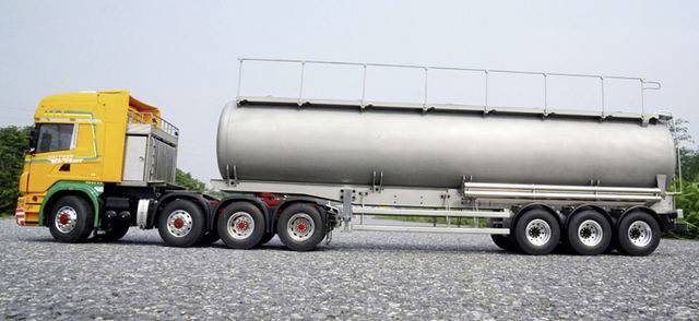 Żółta ciężarówka z srebrną przyczepą cysternową stoi na brukowanej drodze. W tle widoczne są drzewa.