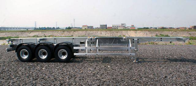 Trzyosiowa rama przyczepy na żwirowym podłożu, bez zabudowy lub ładunku. W tle widoczny jest płaski krajobraz z budynkami.