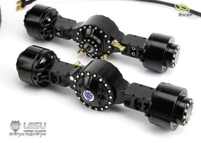 Dwa czarne elementy osiowe do modelarstwa, z widocznymi śrubami i kablami, leżące na białej powierzchni.