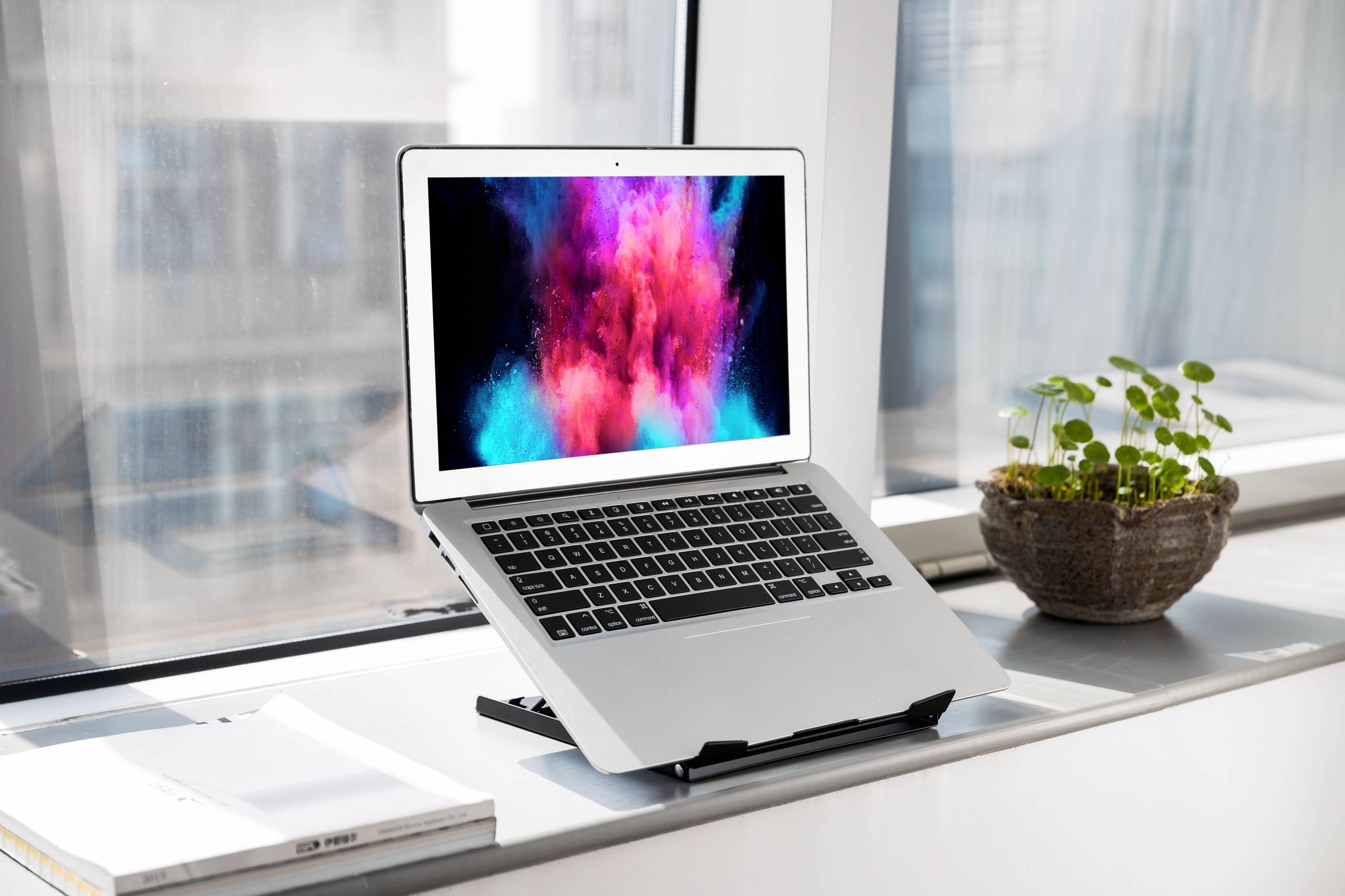 Laptop na parapecie wyświetlający kolorowy ekran. Obok rośliny i notes. Nowoczesne, jasne środowisko pracy.