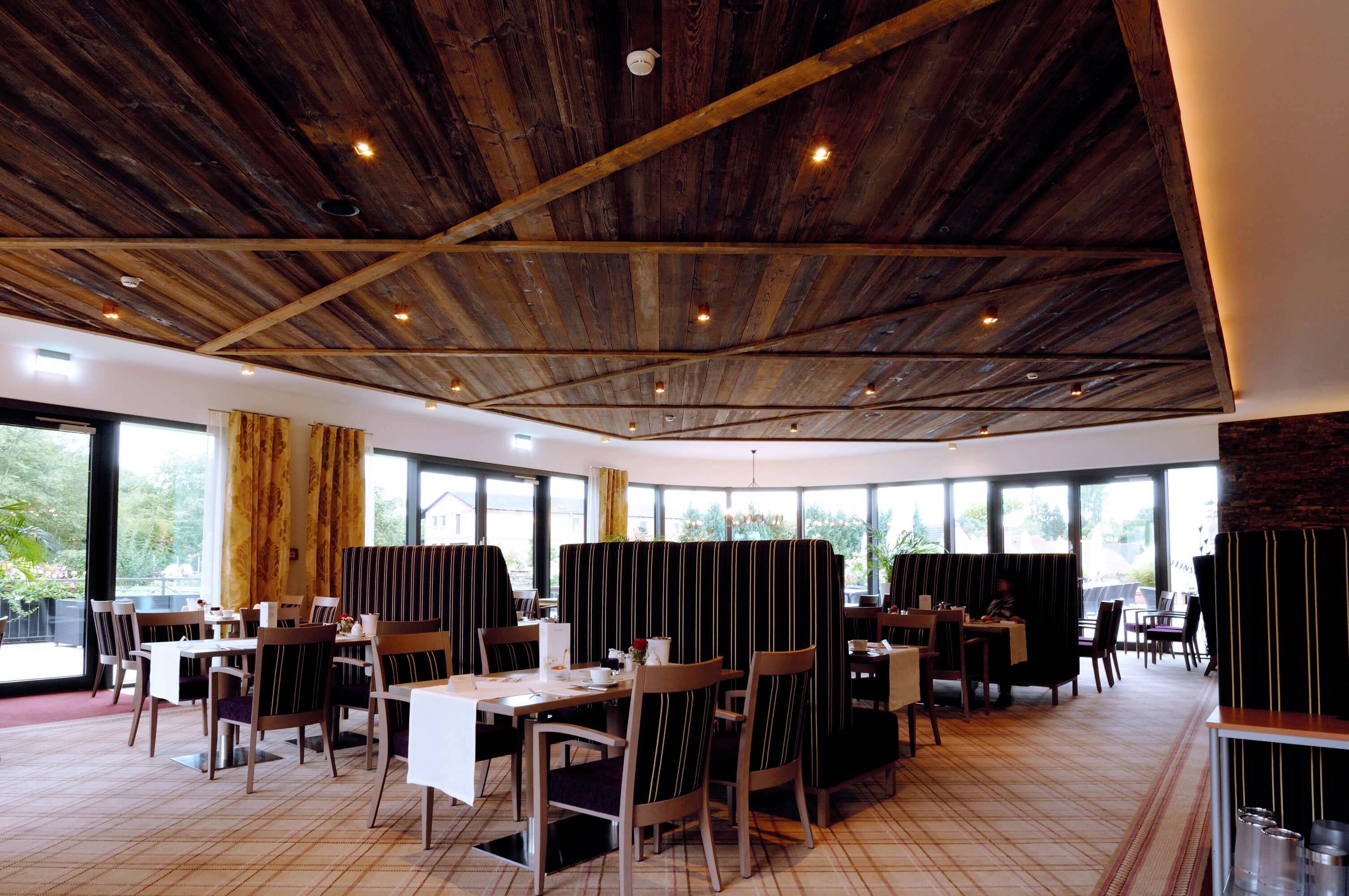 Nowoczesna restauracja z elementami drewnianymi na suficie i ścianach, pustymi stolikami i wyściełanymi strefami siedzącymi, dużymi oknami z widokiem na外trze.