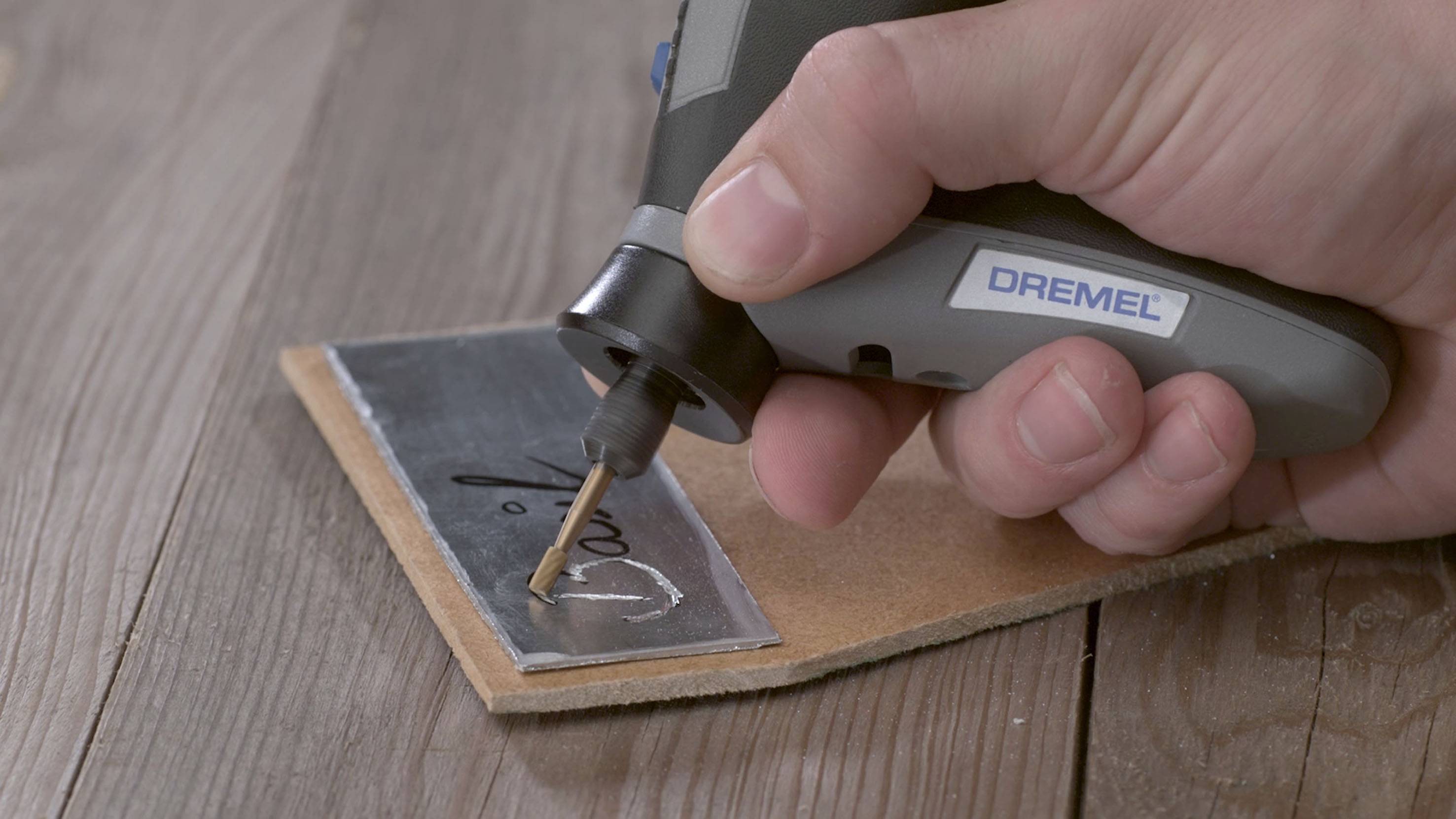 Jedna ręka trzyma elektryczne narzędzie do grawerowania, którym wgrywa imię 'Dawid' w metalową tabliczkę leżącą na drewnianym stole.