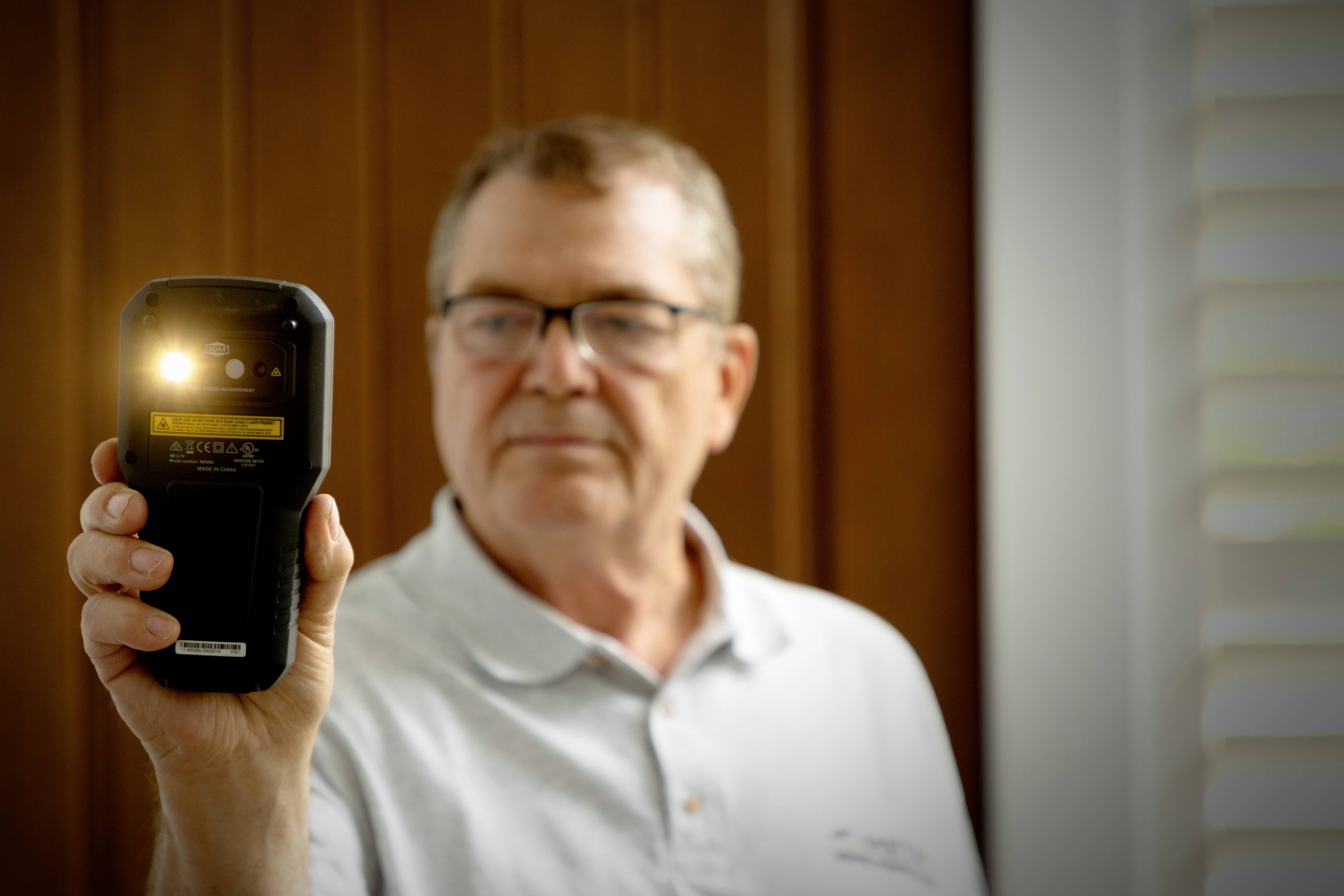 Mężczyzna trzyma miernik natężenia światła i mierzy oświetlenie w pomieszczeniu. Ma na sobie jasną koszulę i stoi przed drewnianym tłem.