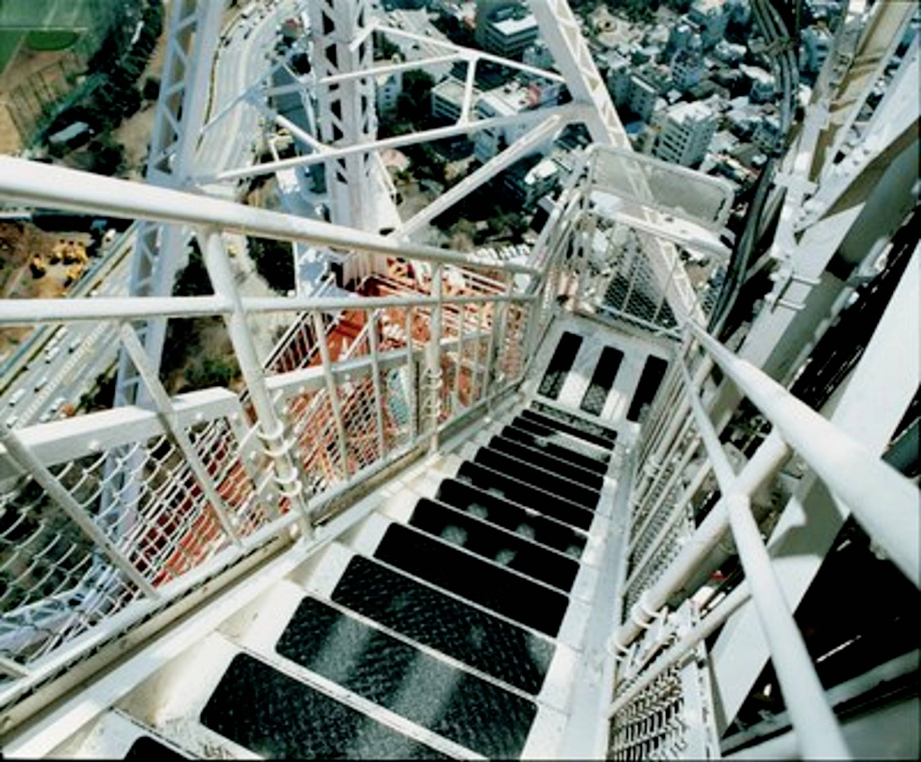 Widok w dół po stromych metalowych schodach na wysokiej strukturze, z panoramą miasta widoczną poniżej, ilustrujący wysokość i środowisko miejskie.