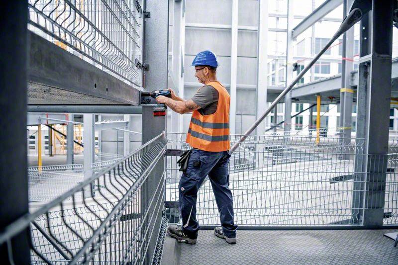 Robotnik budowlany w kasku i kamizelce odblaskowej pracuje narzędziem przy stalowej konstrukcji na terenie budowy.