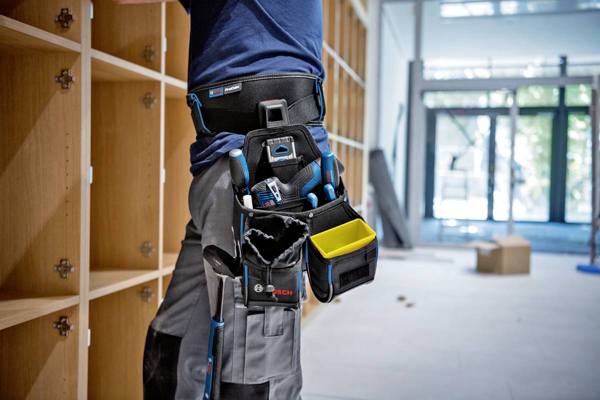 Osoba pracuje w pustym pokoju z regałami na książki, nosząc pas narzędziowy z różnymi narzędziami i akcesoriami.