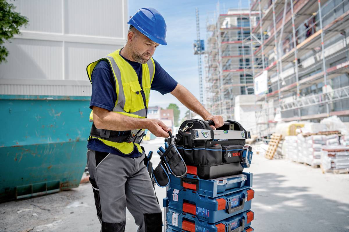 Robotnik budowlany w niebieskim kasku i kamizelce ochronnej stoi na placu budowy i sprawdza narzędzia w jezdnej skrzynce narzędziowej przed rusztowaniem.