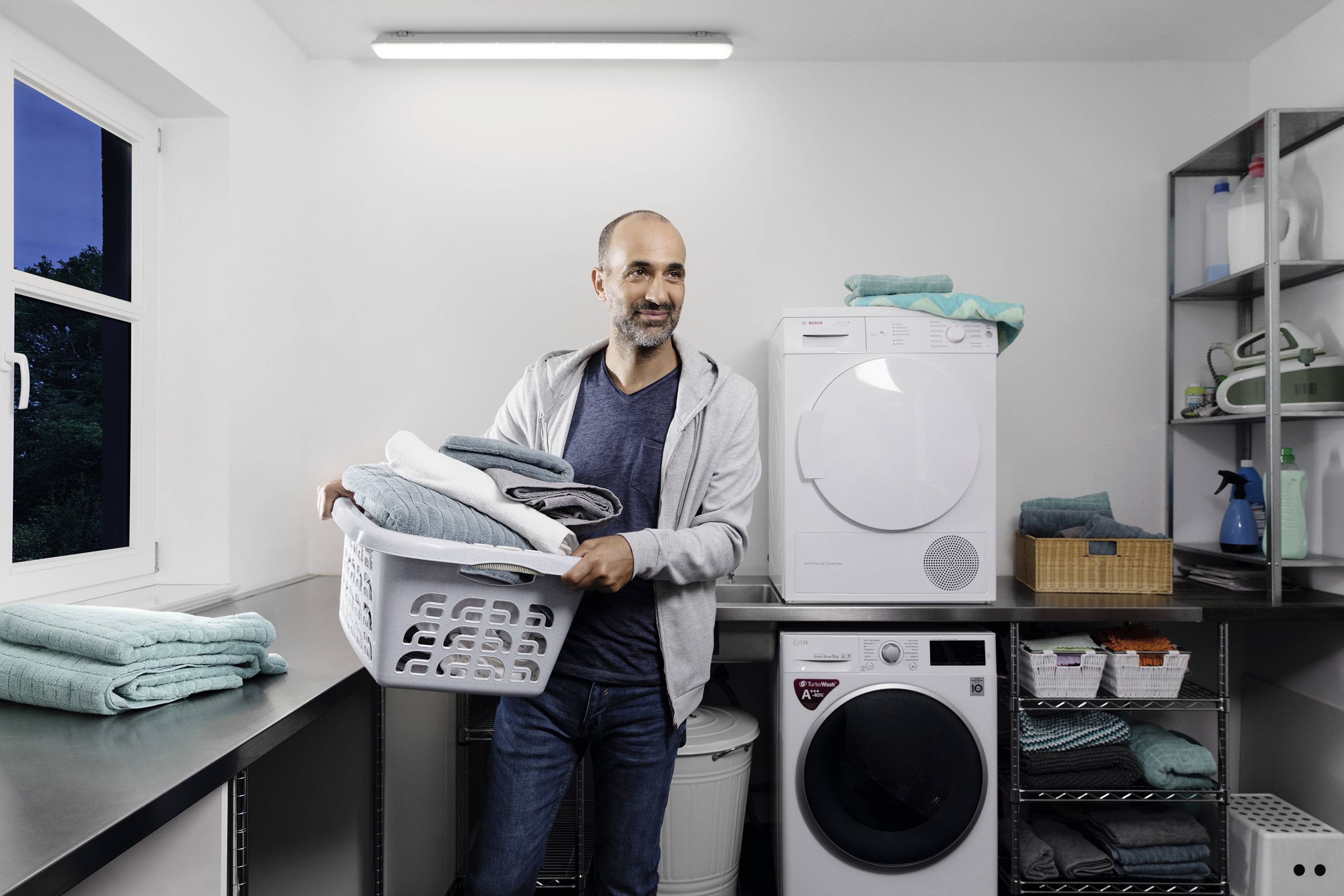 Mężczyzna stoi w pomieszczeniu pralniczym, trzymając kosz pełen starannie złożonych ręczników. W tle widoczna jest pralka i suszarka.