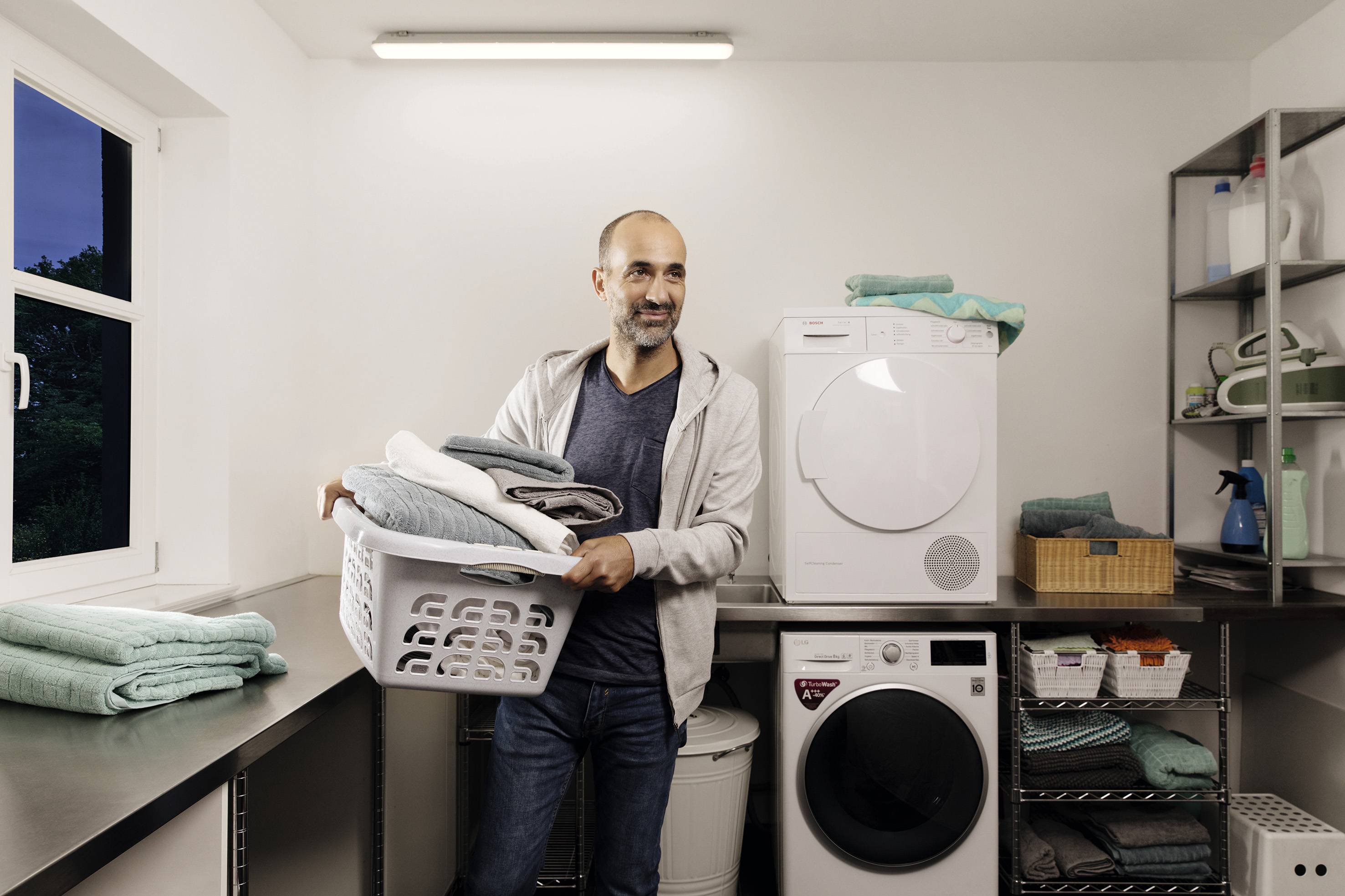 Mężczyzna stoi w pralni z koszykiem pełnym złożonej odzieży. W tle widoczne są suszarka i pralka.