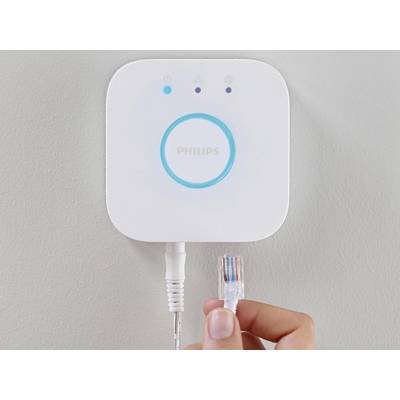 Osoba trzyma wtyczkę sieciową blisko inteligentnej bramki smart home z świecącym niebieskim pierścieniem i logo 'Philips'.