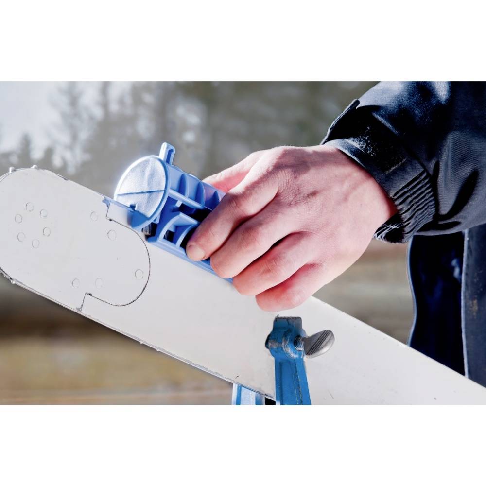 Dłoń trzyma niebieski ostrzacz do narzędzi i kieruje go w stronę piły. Scena rozgrywa się na wolnym powietrzu.