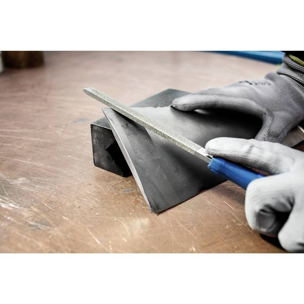 Osoba piluje kawałek metalu pilnikiem. Nosi szare rękawice ochronne, praca odbywa się na stole warsztatowym.