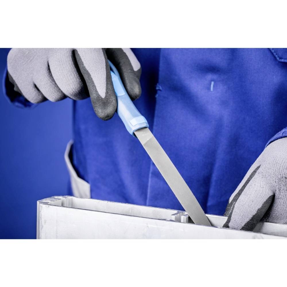 Osoba w niebieskim ubraniu roboczym i szarych rękawicach piluje kawałek metalu pilnikiem.