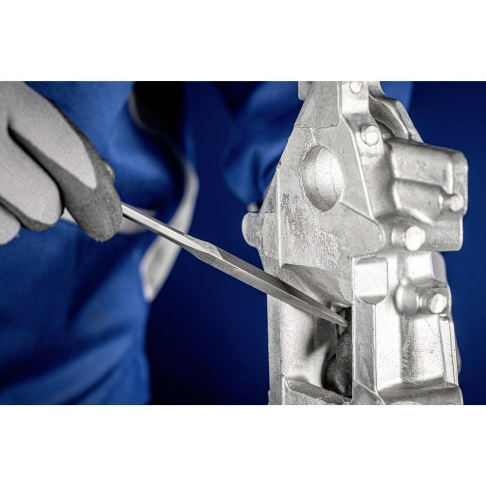 Osoba pilnikuje srebrny element metalowy, ubrana jest w niebieskie ubranie i szare rękawiczki na niebieskim tle.