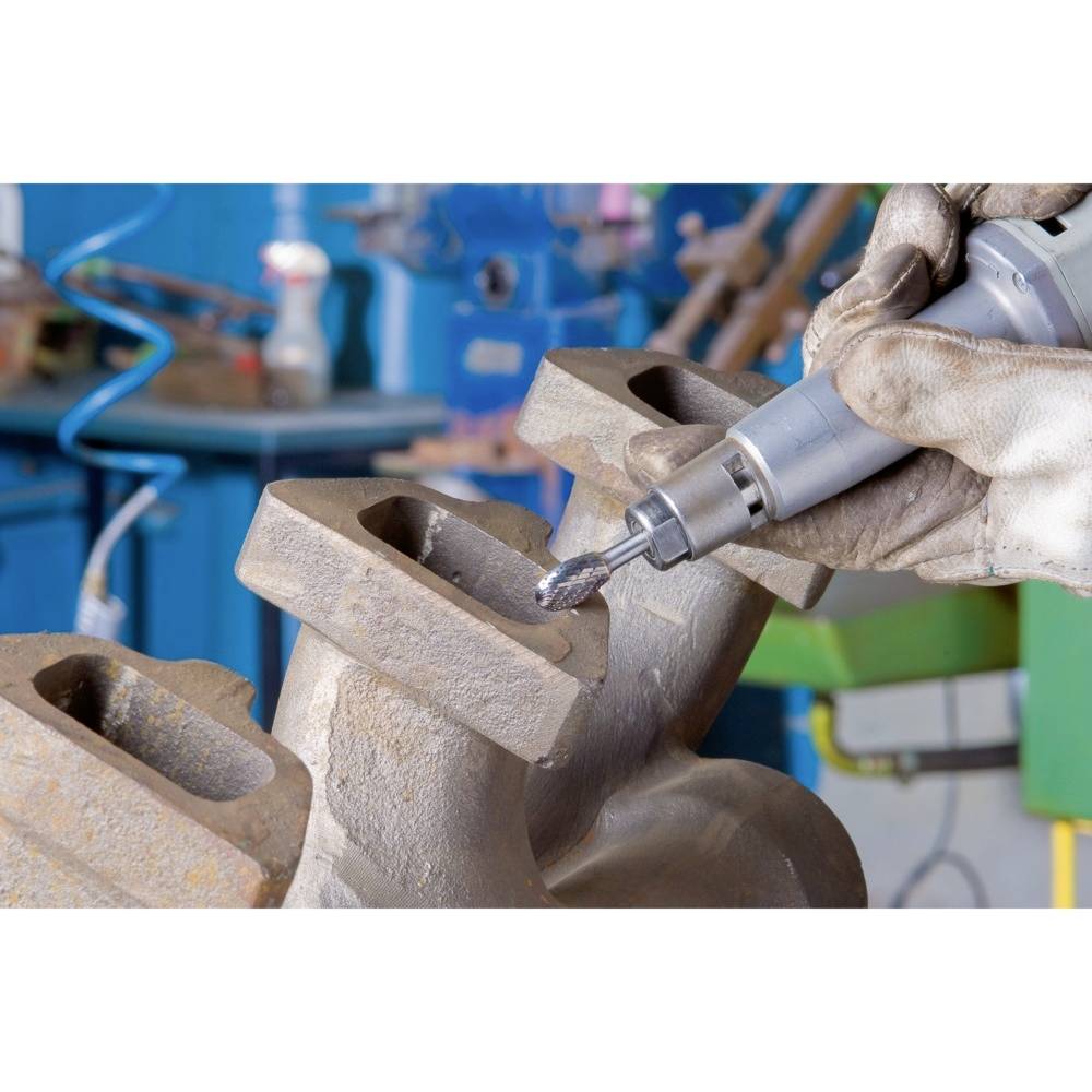 Osoba szlifuje blok metalowy narzędziem ściernym w warsztacie. W tle widać rozmyte wyposażenie warsztatu.