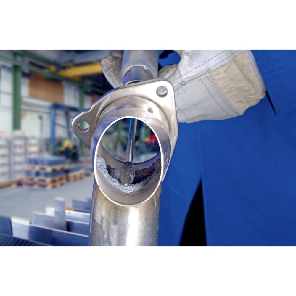 Osoba w ubraniu roboczym obrabia rurę metalową w środowisku przemysłowym. W tle widoczne są regały i sprzęt.