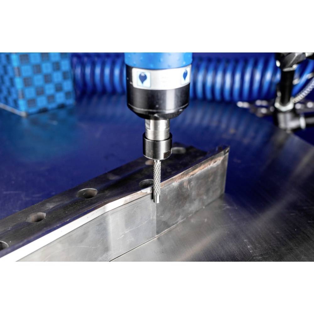 Kawałek metalu jest wiercony przez maszynę. Otoczenie prezentuje elementy przemysłowe, które wskazują na środowisko warsztatu.