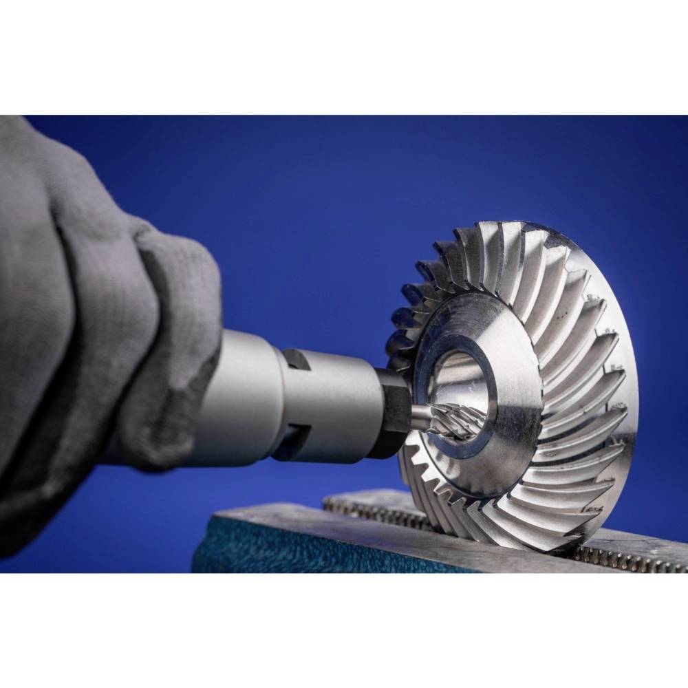 Wiertło obrabia metalową turbinę. Dłoń w szarym rękawicu trzyma urządzenie. Błękitne tło.