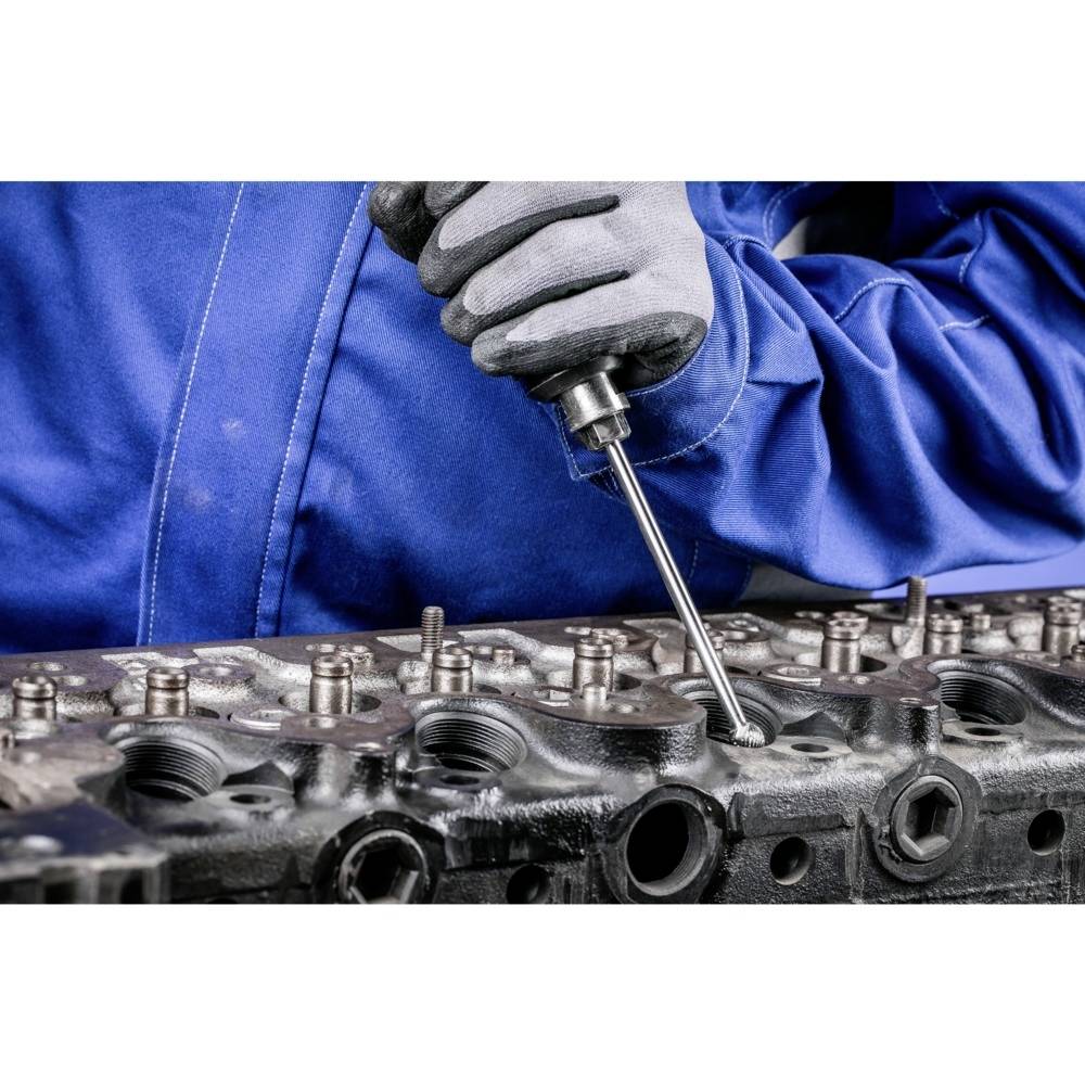 Mechanik w niebieskim kombinezonie roboczym pracuje narzędziem przy części silnika, skupiony na precyzji wykonywanych czynności.