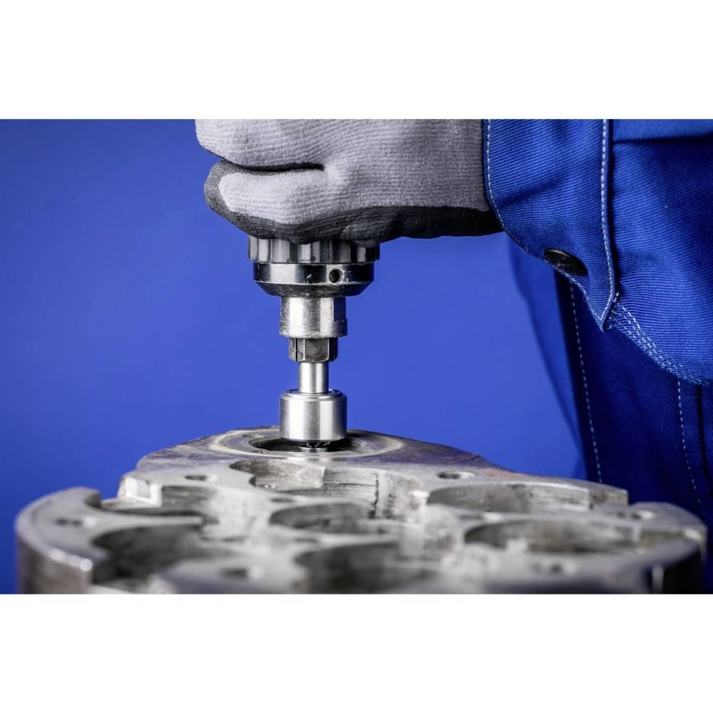 Mechanik pracuje nad powierzchnią metalową, używając wiertła. Osoba ma na sobie rękawice robocze i niebieskie ubranie robocze.