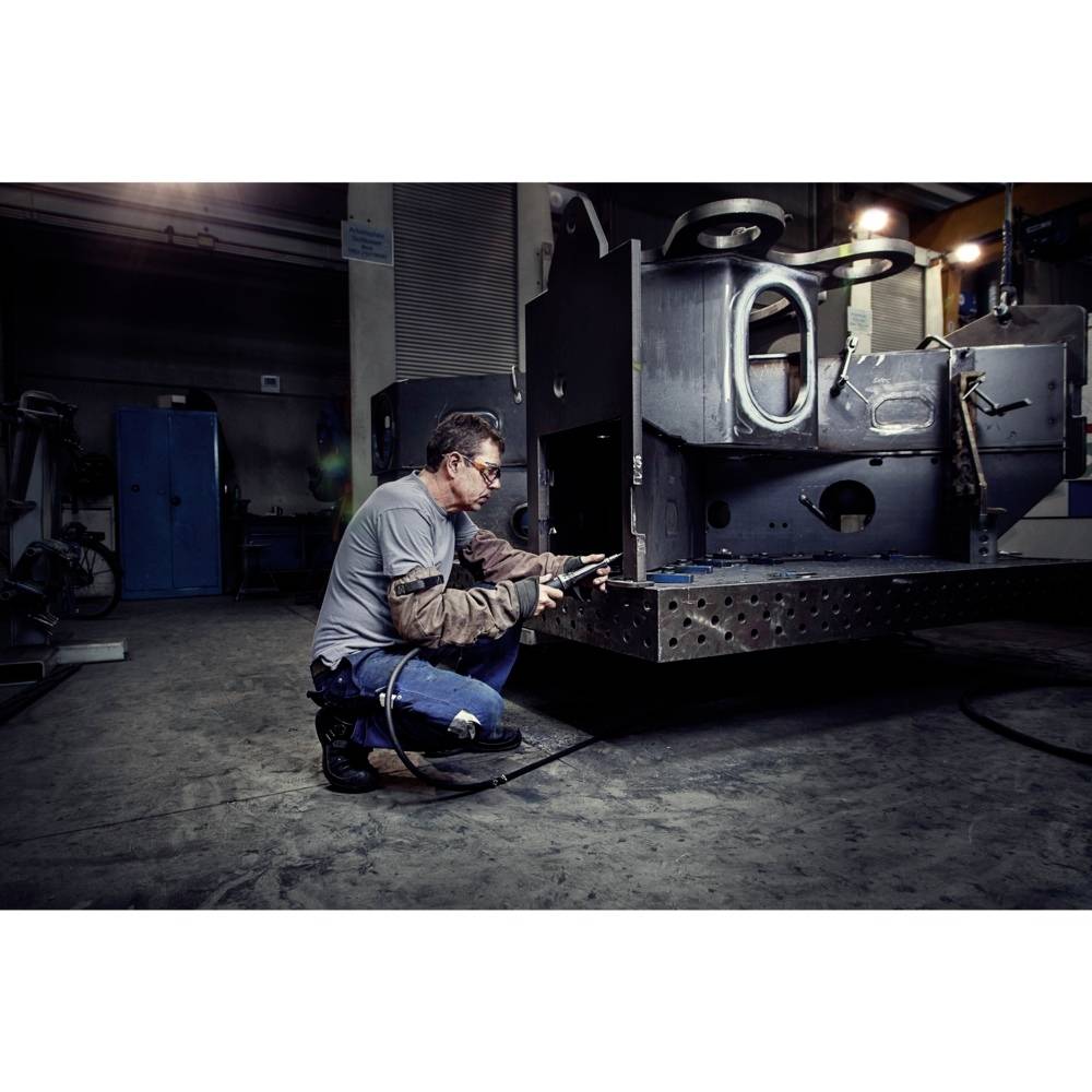 Mężczyzna pracuje z dużym skupieniem, używając narzędzia przy dużym urządzeniu metalowym w przemysłowym środowisku warsztatu.