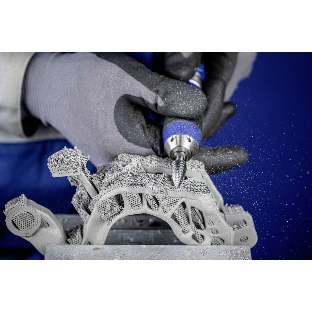 Robotnik w szarych rękawicach obrabia część metalową wydrukowaną w technologii 3D niebieskim narzędziem, a wokół niego lecą iskry.