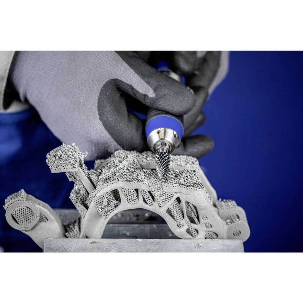 Osoba w odzieży ochronnej obrabia skomplikowany element metalowy za pomocą narzędzia obrotowego. Element charakteryzuje się subtelną, delikatną strukturą.