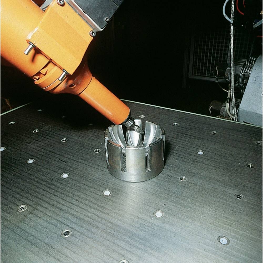 Pomarańczowe ramię robota obrabia cylindryczny element metalowy na stole, w tle otoczone innymi maszynami.
