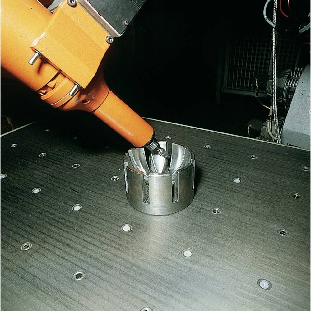 Ramię robota przemysłowego obrabia element metalowy na stole warsztatowym; proces produkcyjny w środowisku warsztatu; praca precyzyjna.