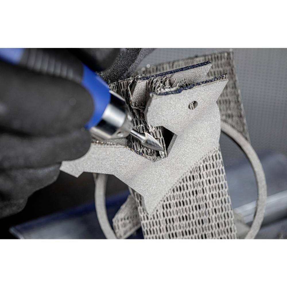 Ręka obrabia szare, strukturalne tworzywo niebieskim narzędziem. Szczegóły materiału są wyraźnie widoczne.