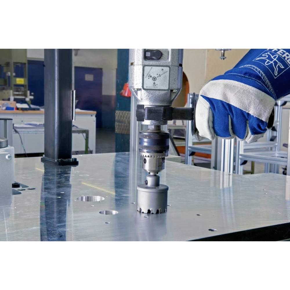 Wiertło pracuje na płycie metalowej. Osoba w niebieskich rękawicach ochronnych trzyma narzędzie. W tle widoczne są stoły warsztatowe.