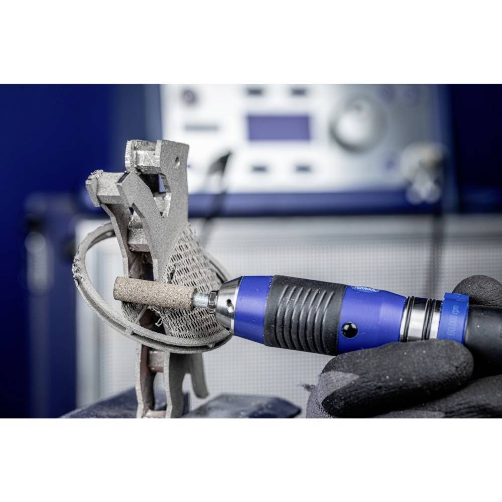 Dłoń trzyma obracające się narzędzie szlifierskie, które obrabia metalowy element wydrukowany w technologii 3D. Na rozmytym tle widać urządzenie techniczne.