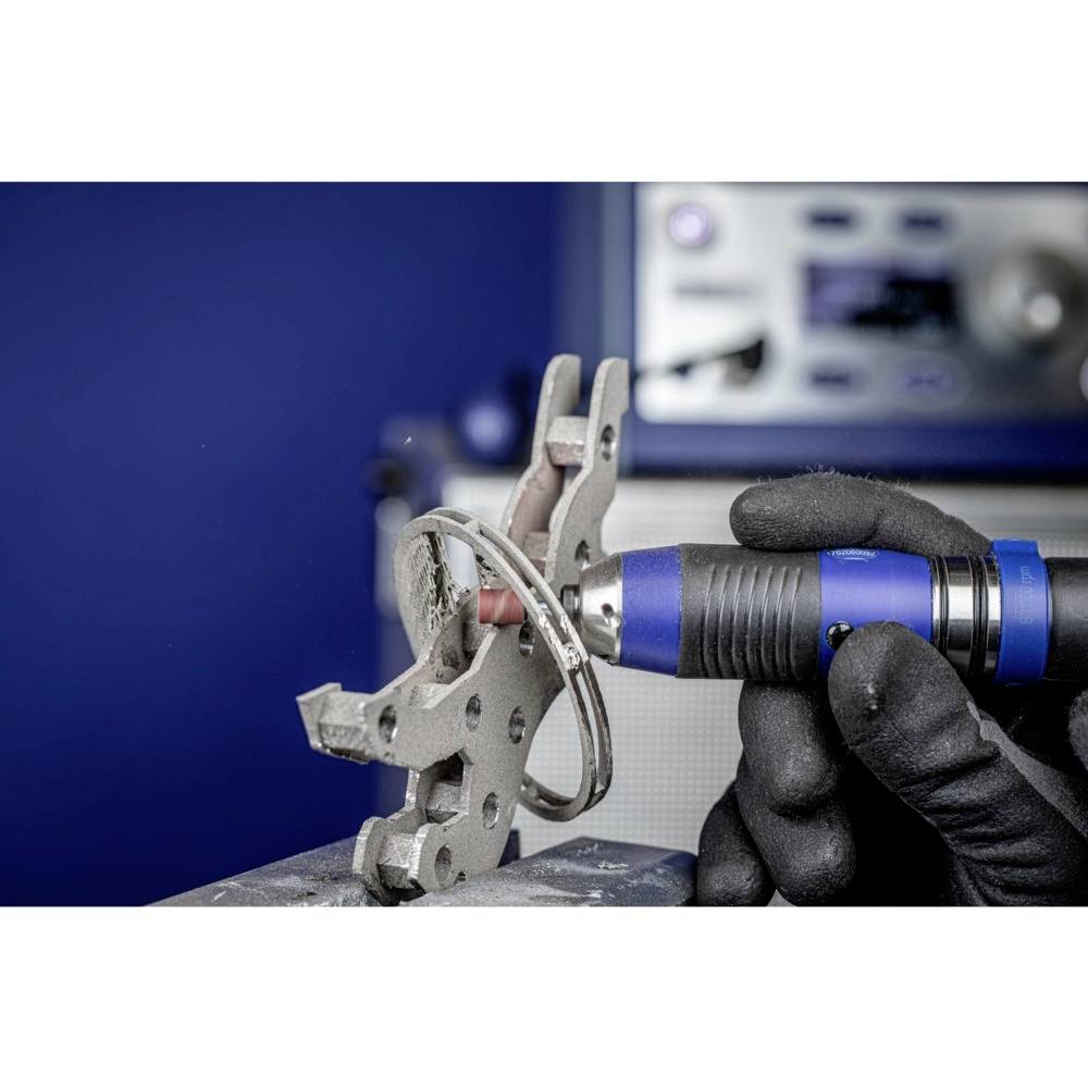 Osoba obrabia element metalowy niebieskim narzędziem szlifierskim w warsztacie. Na tle widać rozmazane urządzenie.