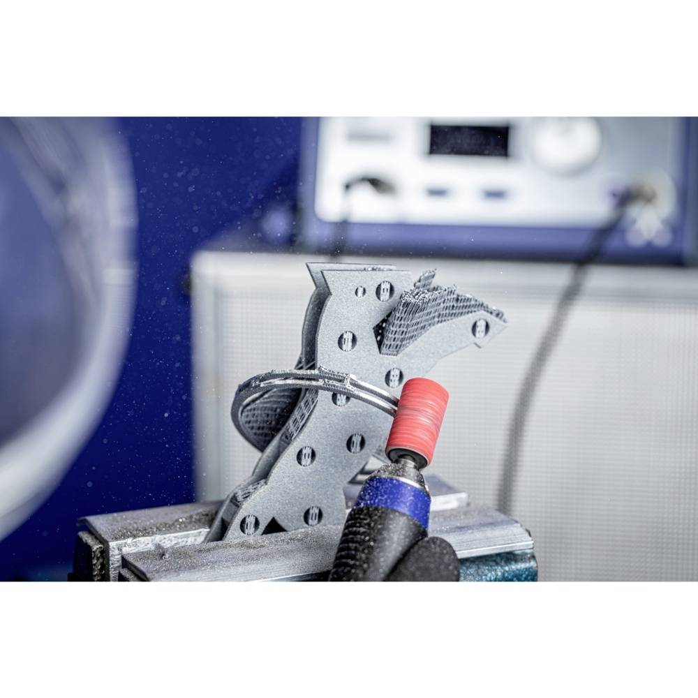 Osoba poleruje element przemysłowy za pomocą narzędzia obrotowego, aby usunąć wady powierzchni. Na tle widoczne są urządzenia techniczne.