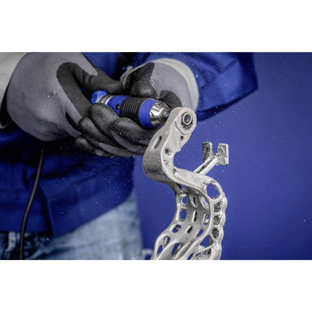 Osoba w rękawicach obsługuje elektronarzędzie do szlifowania kawałka metalu. Na tle niebieskiej ściany.