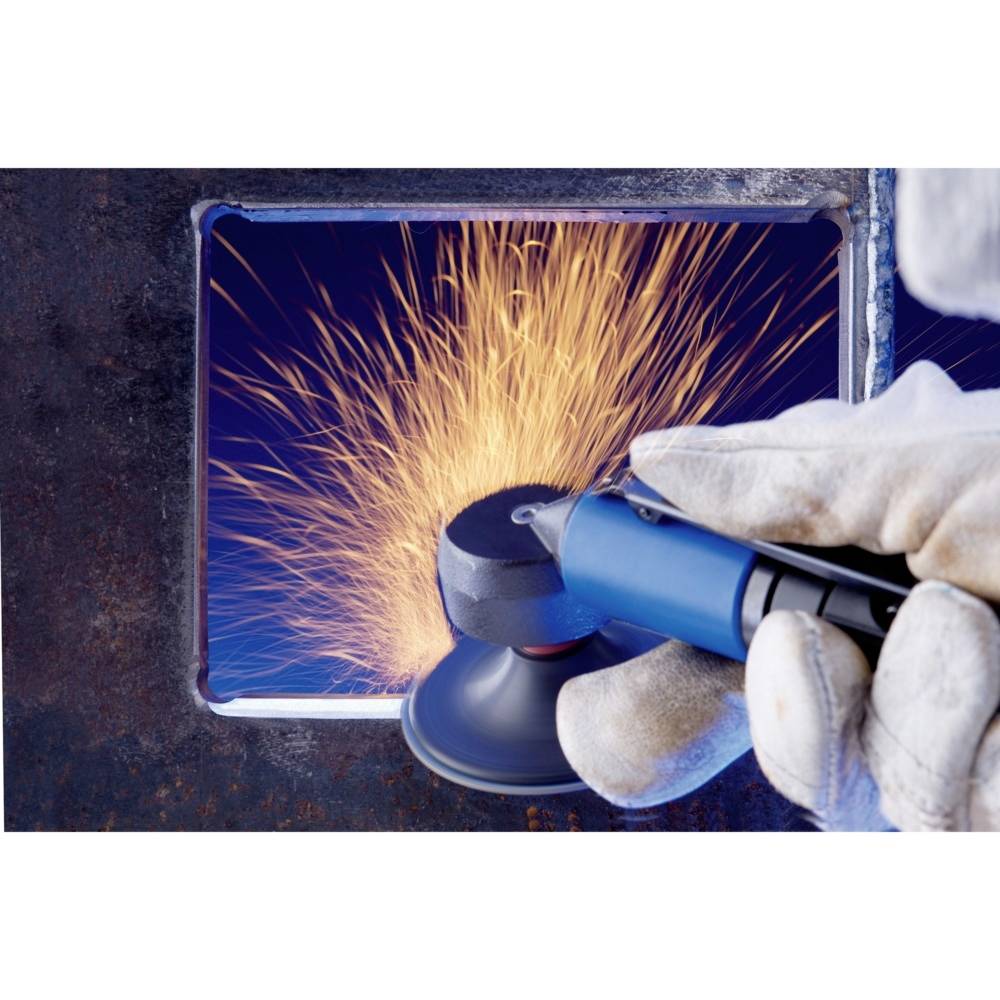 Robotnik w rękawicach trzyma narzędzie, podczas gdy za małym okienkiem w metalowym panelu tryskają iskry.