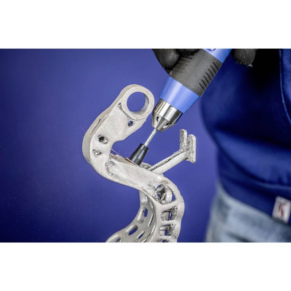 Osoba poleruje metalową część wydrukowaną w technologii 3D ręcznie prowadzonym narzędziem. Tło jest niebieskie.