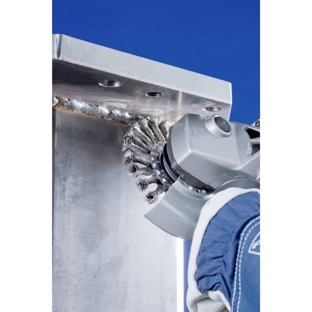 Technik szlifuje krawędź metalową za pomocą szlifierki wyposażonej w szczotkę. W tle widać błękitne niebo.