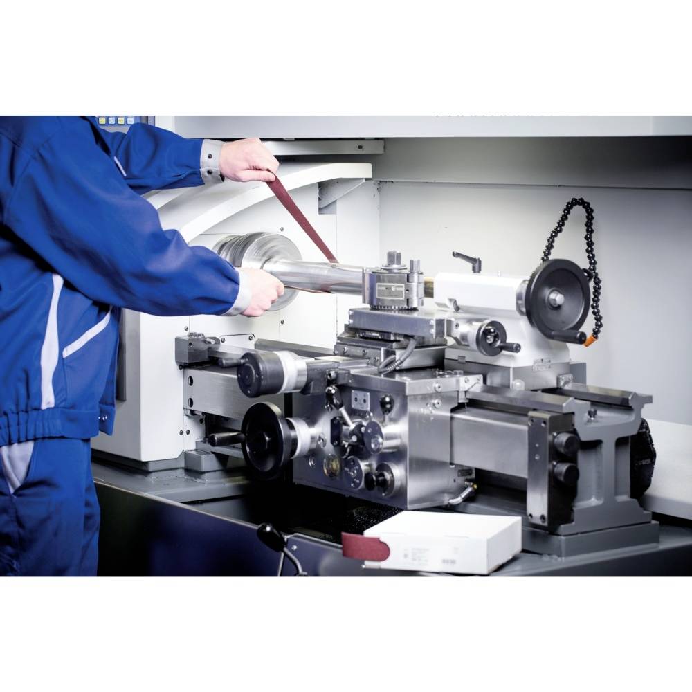 Technik pracuje przy tokarce i obrabia element metalowy za pomocą taśmy szlifierskiej. Maszyna jest srebrzysta i skomplikowana.