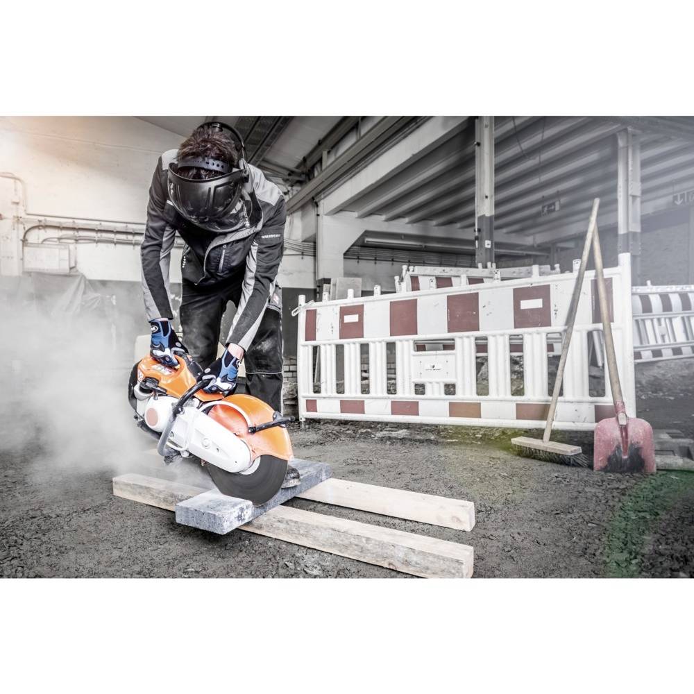 Robotnik budowlany przecina beton zmotoryzowaną tarczą tnącą, otoczony barierkami ochronnymi i pyłem.