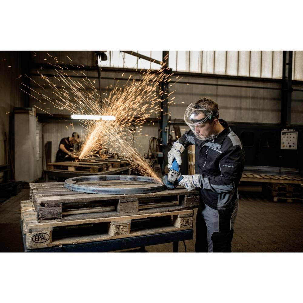 Pracownik w odzieży ochronnej szlifuje metal, powodując lot iskier w hali warsztatowej, a w tle widoczni są kolejni pracownicy przy stanowiskach roboczych.