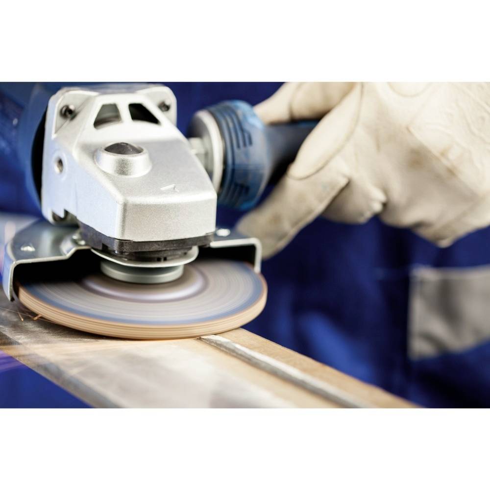 Robotnik szlifuje kawałek metalu za pomocą szlifierki kątowej. Szlifierka jest w ruchu, co widać po obracającej się tarczy.