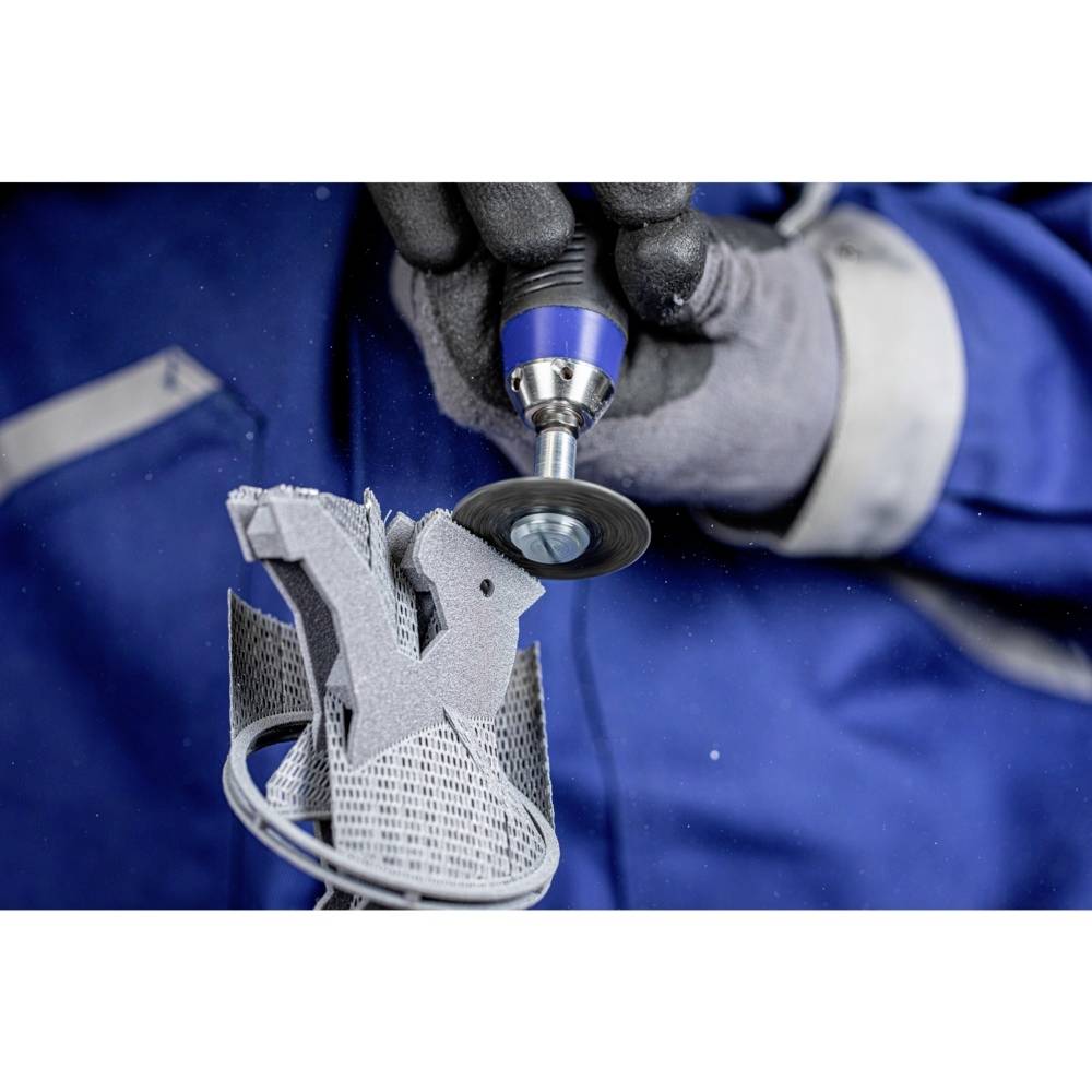 Osoba ubrana w niebieskie ubranie robocze obrabia narzędziem elektrycznym część metalową wydrukowaną w technologii 3D. Dookoła unoszą się drobne wióry metalowe.