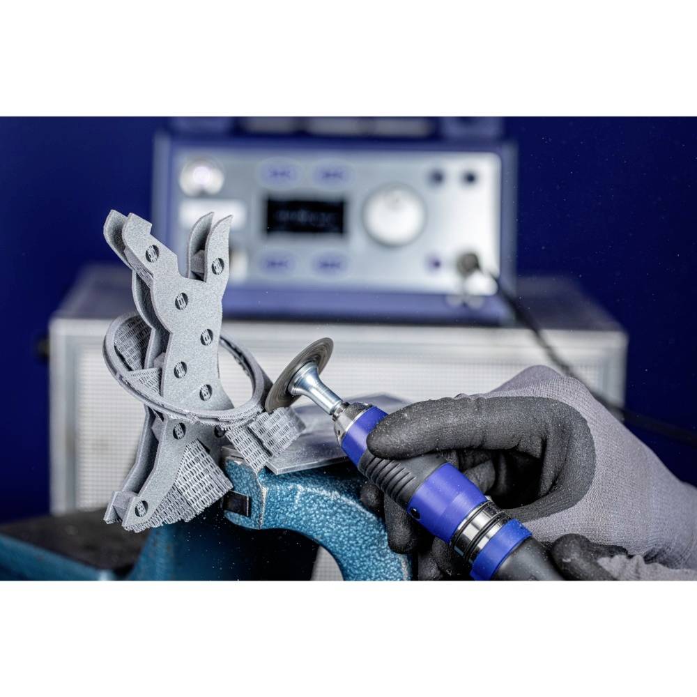 Ręka w rękawicy obsługuje szary model elektronarzędziem. Na tle widać urządzenie z wyświetlaczami i przyciskami.