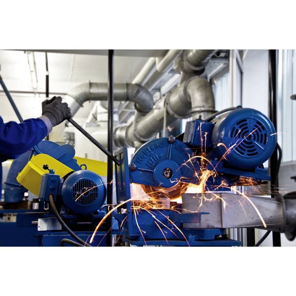 Robotnik w odzieży ochronnej obsługuje przemysłową szlifierkę, z której tryskają iskry. Otoczenie wypełnione dużymi przewodami wentylacyjnymi.