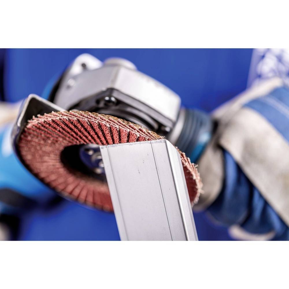 Robotnik szlifuje kawałek metalu za pomocą obracającej się tarczy szlifierskiej, ubrany jest w niebieskie rękawice ochronne i niebieski strój roboczy.