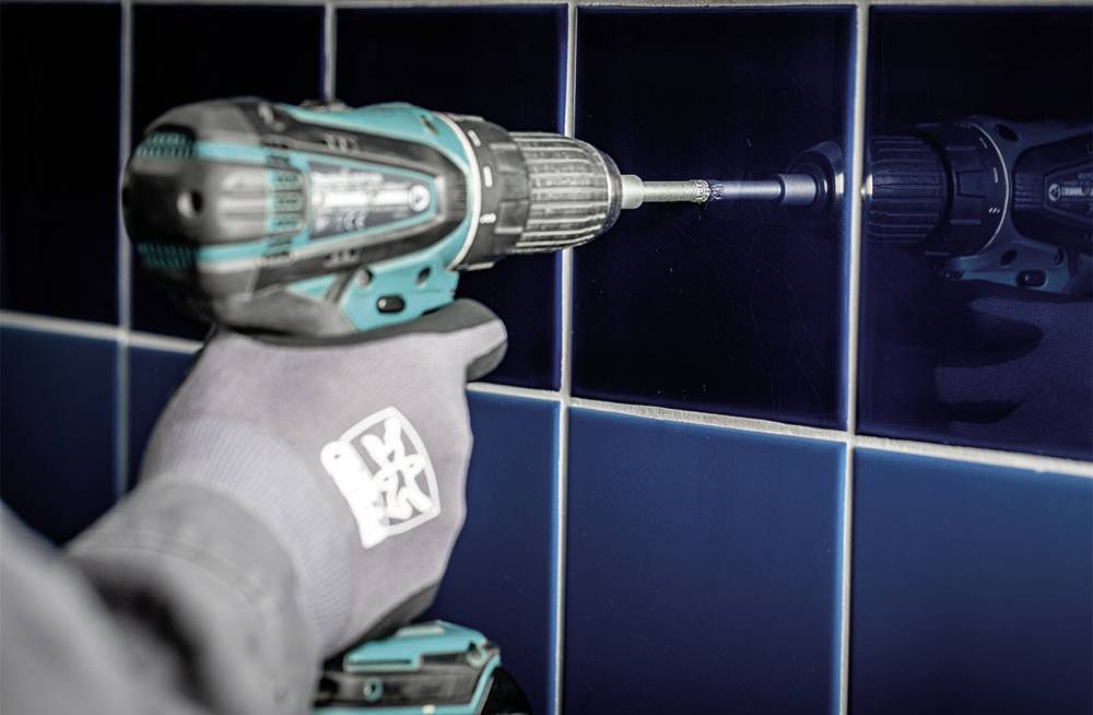 Dłoń w rękawicy trzyma akumulatorowy wkrętak, który wwierca się w niebieskie kafelkowane ścianę.